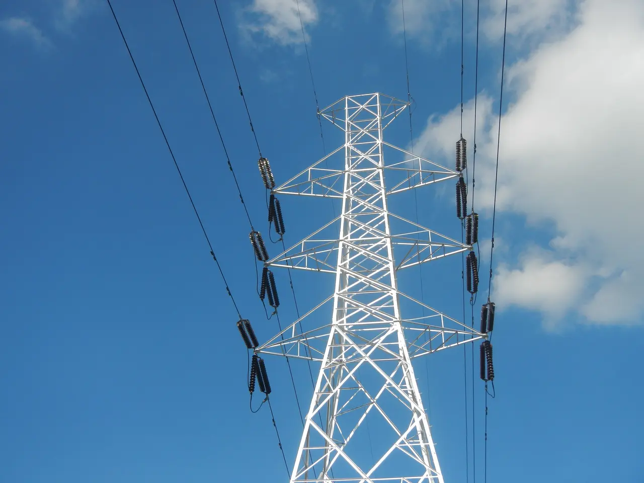 The image shows a high voltage power line against a blue sky with white, fluffy clouds. The pylon...