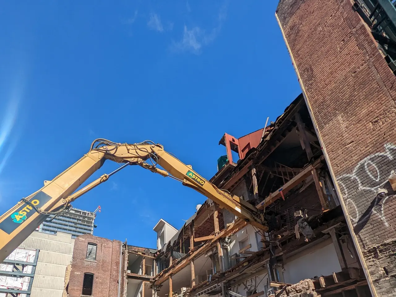 The image shows an excavator demolishing a building in the middle of a city, with a crane in the...