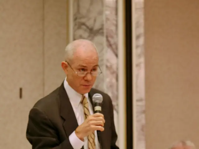 This man wore suit and tie is holding a mic and paper. In-front of this man there is a table, on a...