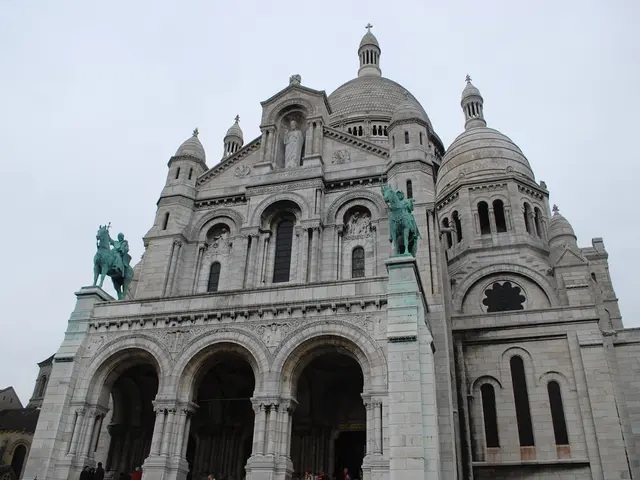 In this picture there is a monument in the center of the image, there are houses and a tree in the...
