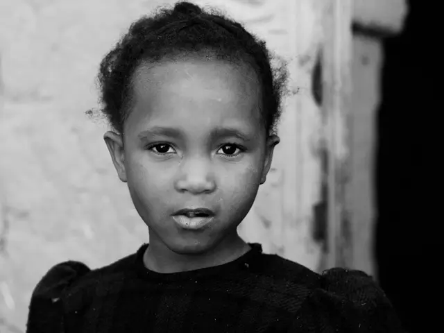 In this black and white image, we can see a kid wearing clothes. In the background, image is...