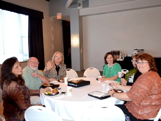 This is the picture of a room. In this image there are group of people sitting around the table....