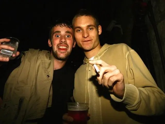 There are two men standing holding glass in one hand and cigarette in another hand.