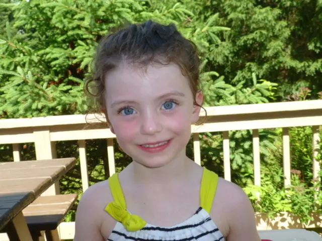 In this image there is a kid wearing white color dress looking at the camera and at the background...