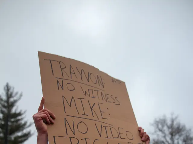 In this image people are protesting on road, a man holding sheet in his hand some text is written...
