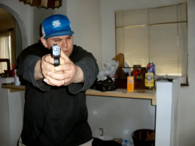 In the image there is a man, he is holding a gun with the both hands and behind him there is a...