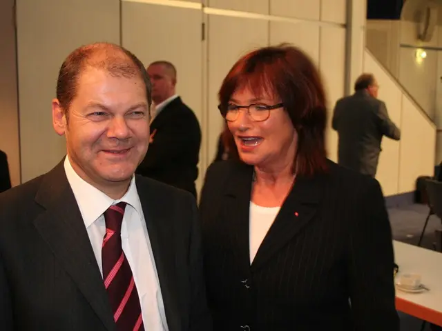 In this picture we can see a man and a woman in black blazers smiling. On the background we can see...