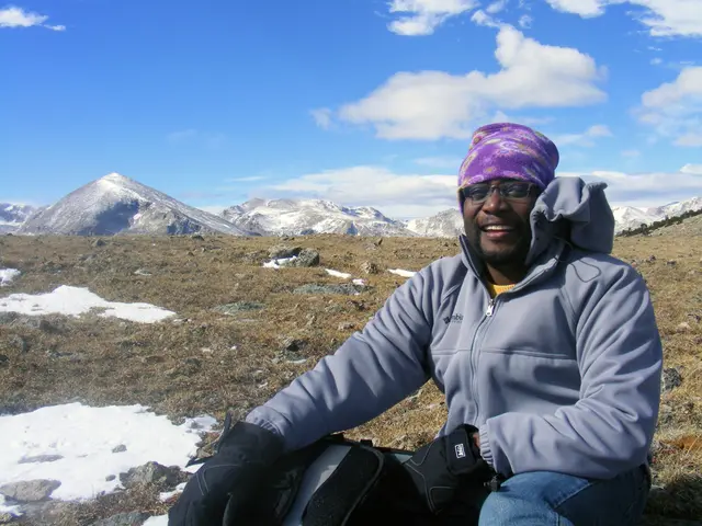 In this image in the front there is a man sitting and smiling. In the background there is dry grass...