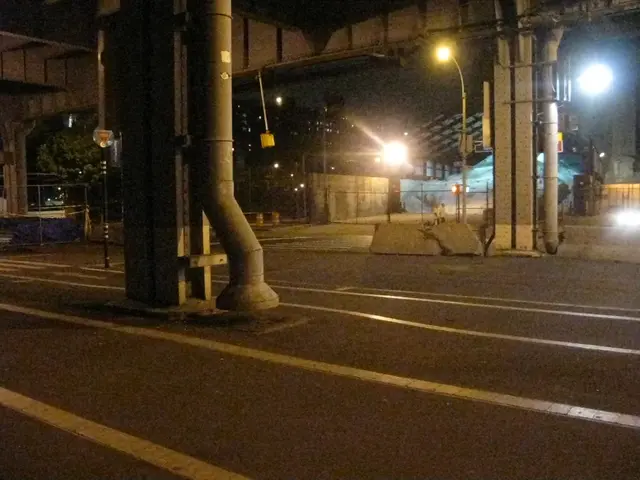 In the foreground of this image, there is a road, pillars, pipes, poles,lights, a shed, fencing,...