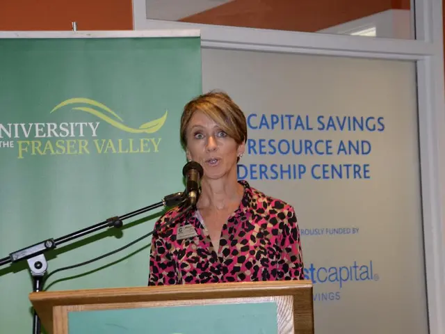 In the center of the image we can see a lady standing and talking. We can see a mic before her. At...