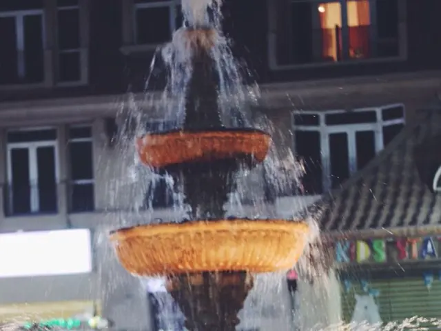 In front of the picture, we see the water fountain. On the right side, we see a building in green...