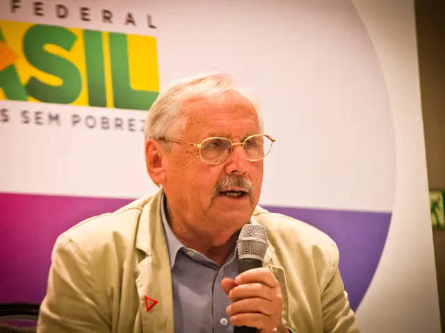 in this image i can see a person speaking, holding a microphone in his hand. behind him there is a...