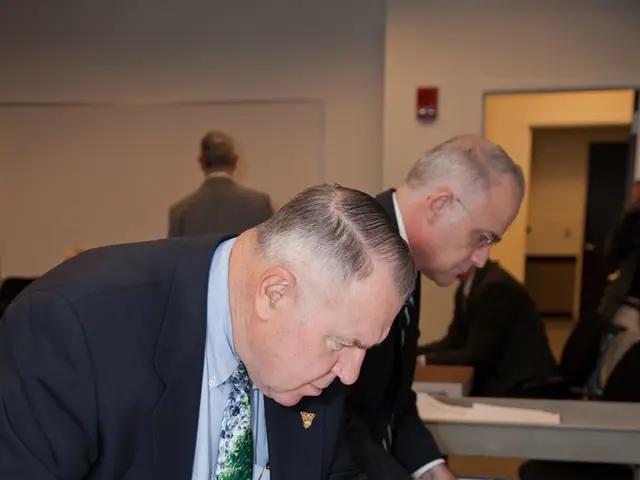 In this image I see 2 men who are wearing suits and I see that this man is holding papers and I see...