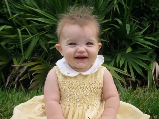 This is a small girl sitting and smiling. I can see the grass. In the background, I think these are...