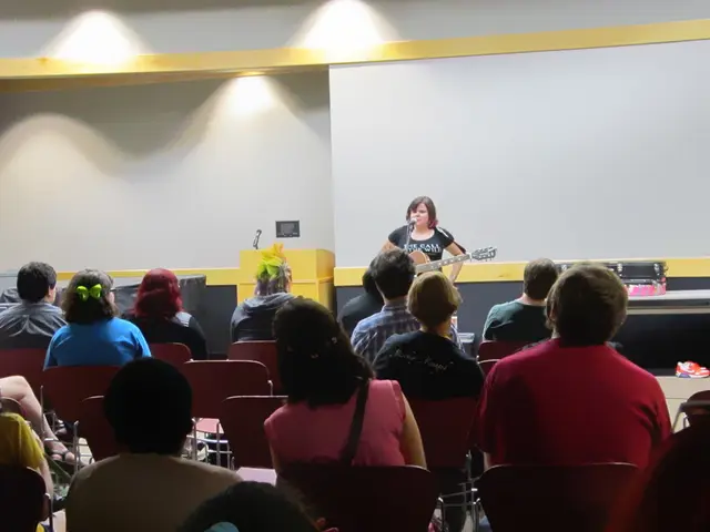 In this image, There are some people sitting on the chairs which are in red color, In the middle...