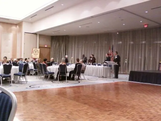 In this picture I can see group of people sitting on the chairs, there are few people standing,...