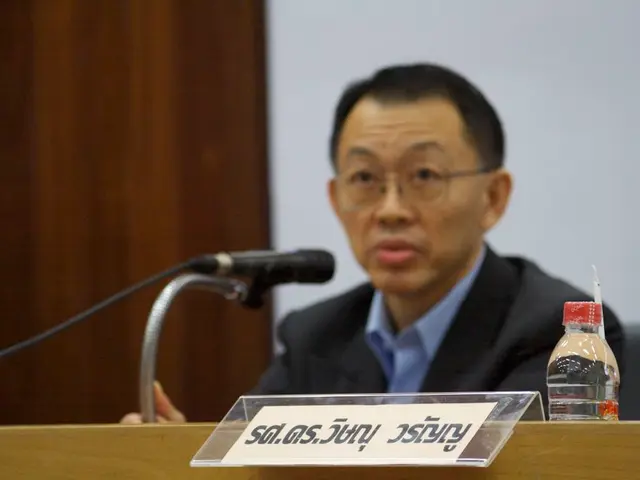 There is a person sitting on a chair. He is wearing a suit and he is speaking on a microphone.