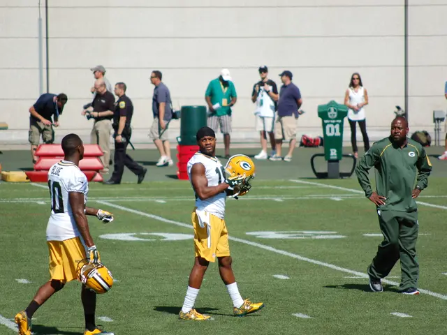 In this image there are two people who are wearing jersey, and they are holding helmets. At the...