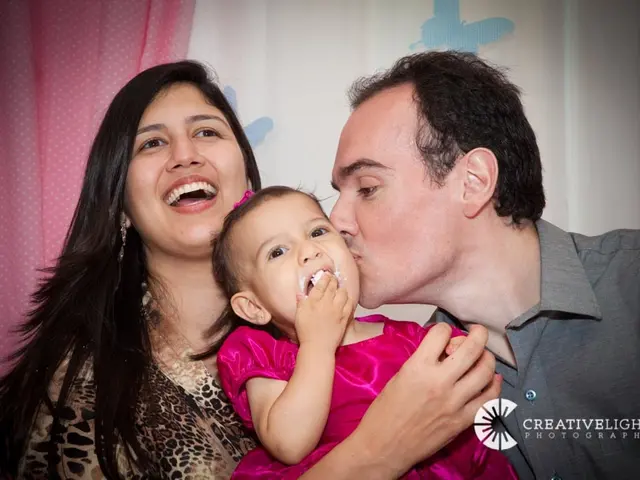 In this image there is one man and one woman a man is holding a baby, in the background there is a...