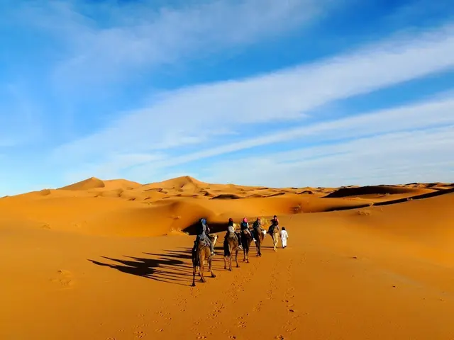 In this image there is a desert, five people riding camels on the desert and a person is walking,...
