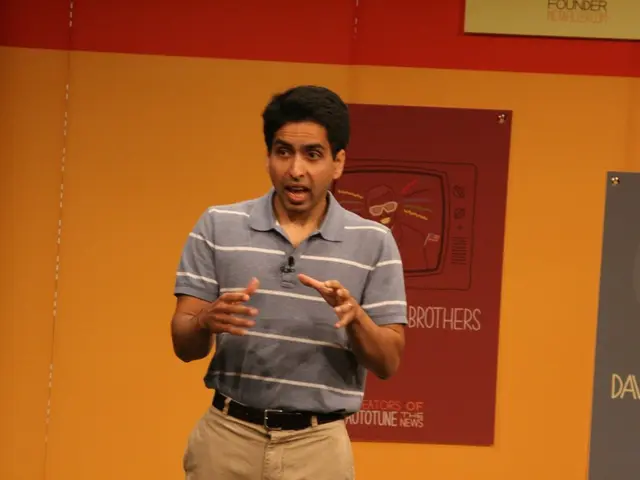 In this image there is a man standing on stage and speaking behind him there is a wall with some...