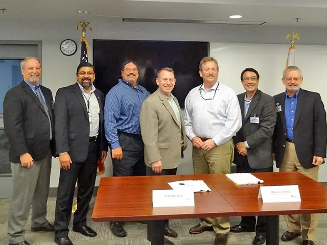 people are standing in a room. in front of them there is a table on which there are papers. behind...