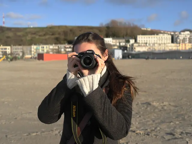 In this image i can see a woman is holding a camera. Behind this woman i can see there are few...