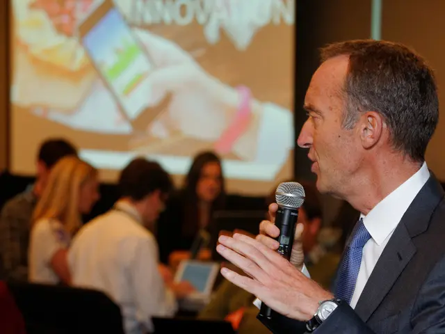 The person wearing suit is speaking in front of a mic and there are few people sitting behind him.