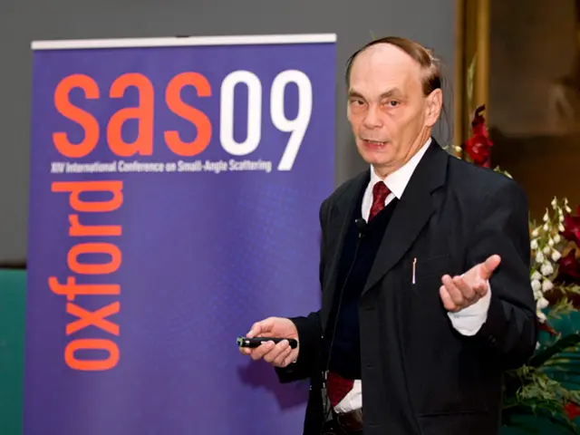 In this image, we can see an old person in a suit is holding an object and talking. Background we...