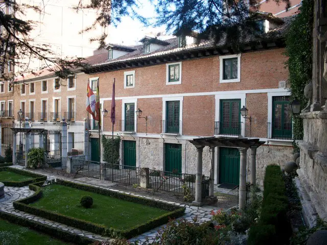 In the picture I can see few buildings and there are few flags,fence and a greenery ground in front...