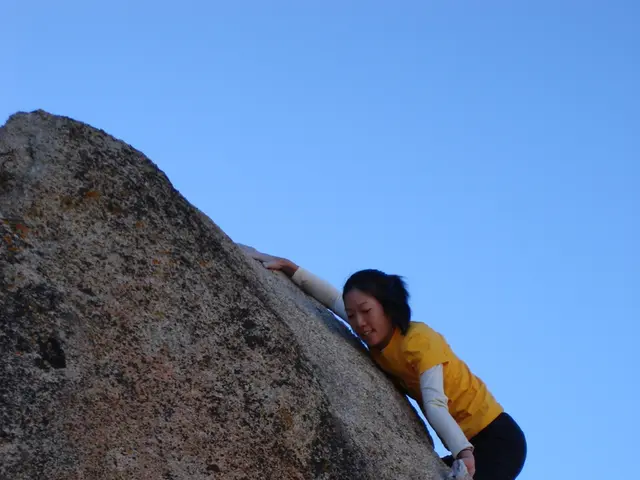 In this image there is a girl climbing a big rock.