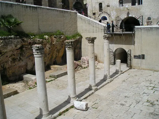 In this image we can see pillars and building. On the stairs of the building people are standing....