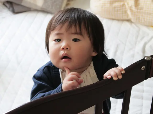 There is a kid standing on the bed, holding one of the railing, with a hand under his chin. He is...