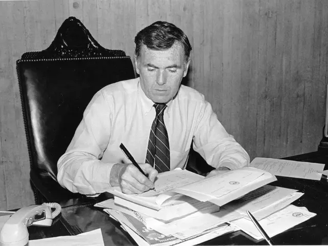 This is black and white picture were a man is sitting on a black chair, he is holding pen. In front...