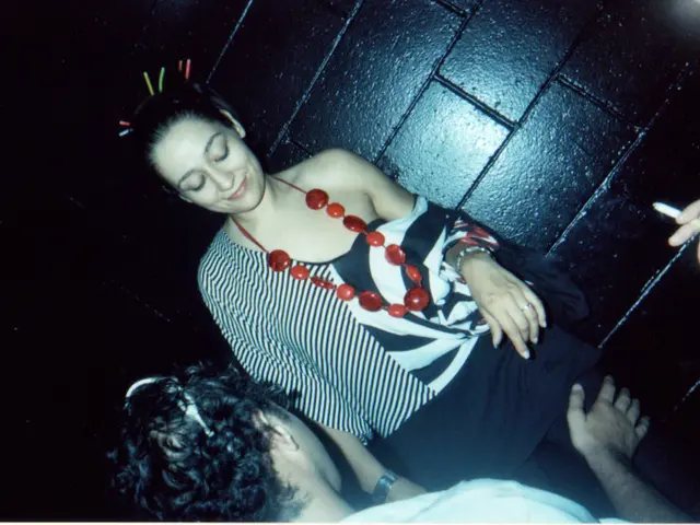 In this image there are two personś, there is a hand holding the cigarette, there is a black wall...