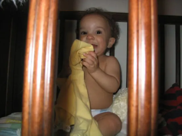 In this image we can see a kid sitting on the bed and holding a cloth and there are few objects...