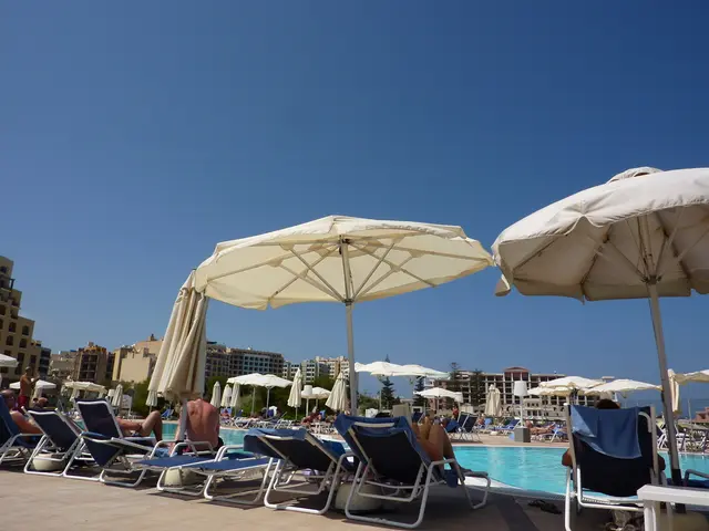In the foreground of this image, there is a beach chairs on which persons are sitting and there are...