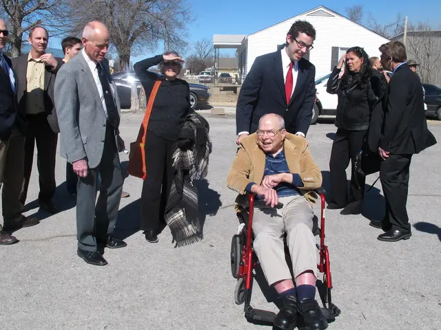 In the center of the image there are people standing. There is a person sitting on wheelchair. In...