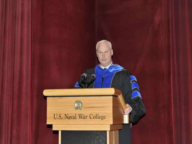 In this image we can see a man standing in front of the podium and also the mikes. In the...