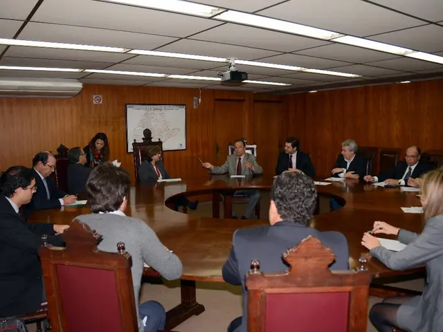 In this image I can see number of people are sitting on chairs. Here I can see a round table. In...