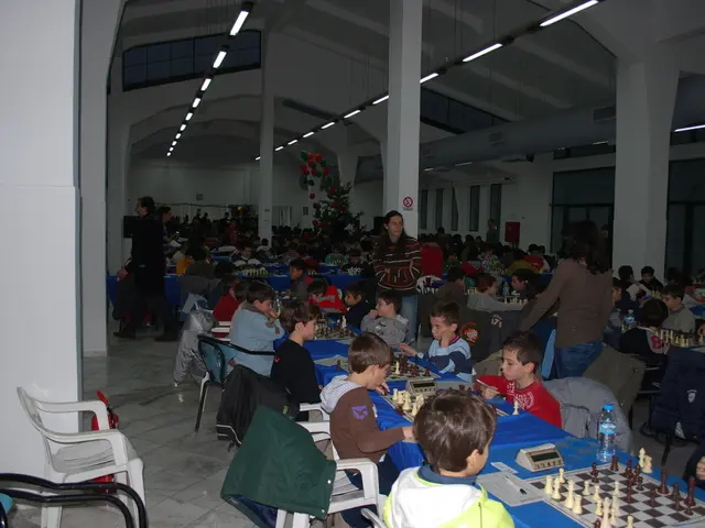 In this image, there are group of people sitting on the chair, in front of table and playing chess...