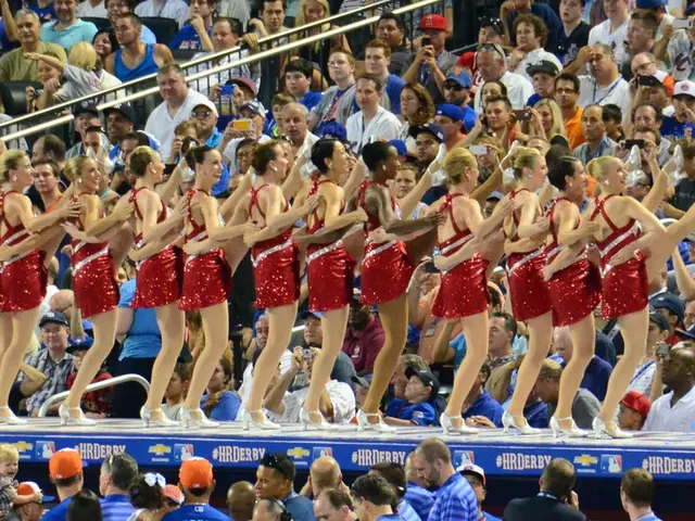 In the center of the image we can see girls dancing. In the background there is crowd sitting.