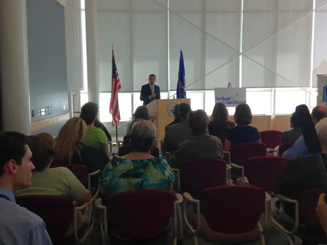 There are many people sitting in the chairs in this room and one guy is standing near the podium...