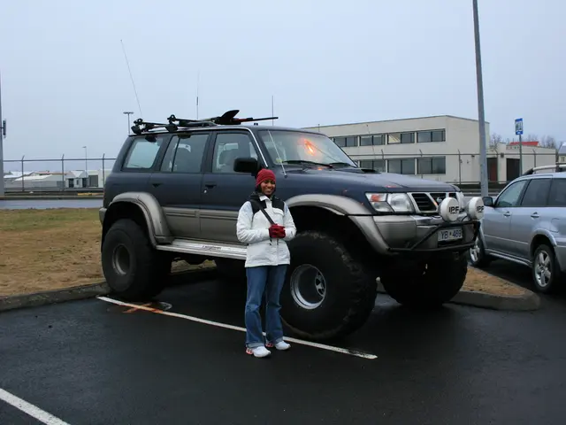 In the image there are two cars and there is a woman standing in front of the first car, behind the...