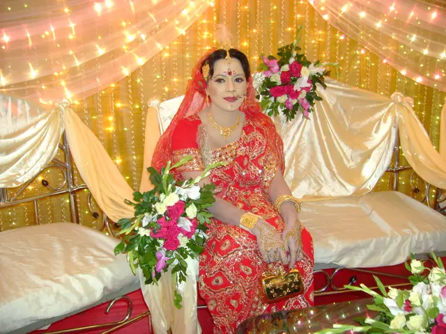In this image, we can see a woman in red saree is sitting on the sofa. Here we can see few flower...