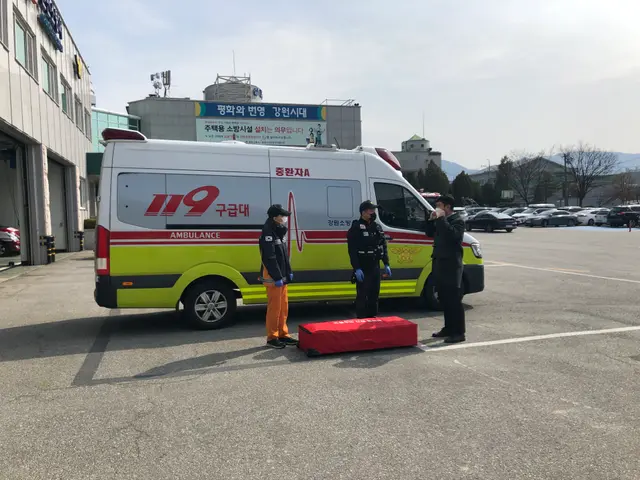 The image shows an ambulance parked in a parking lot next to a building, with three people standing...