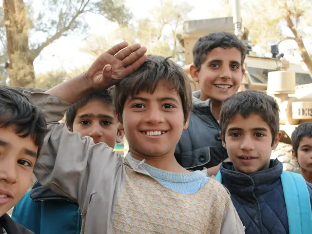 The image shows a group of young boys standing next to each other in front of a military vehicle,...