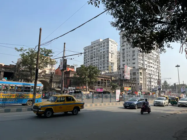 The image shows a yellow taxi cab driving down a busy street in Mumbai, surrounded by tall...