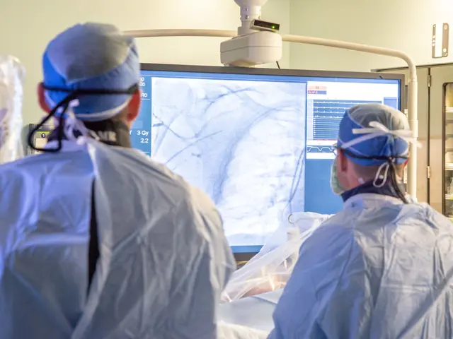 The image shows two surgeons in scrubs and masks looking at an x-ray image on a large screen in an...