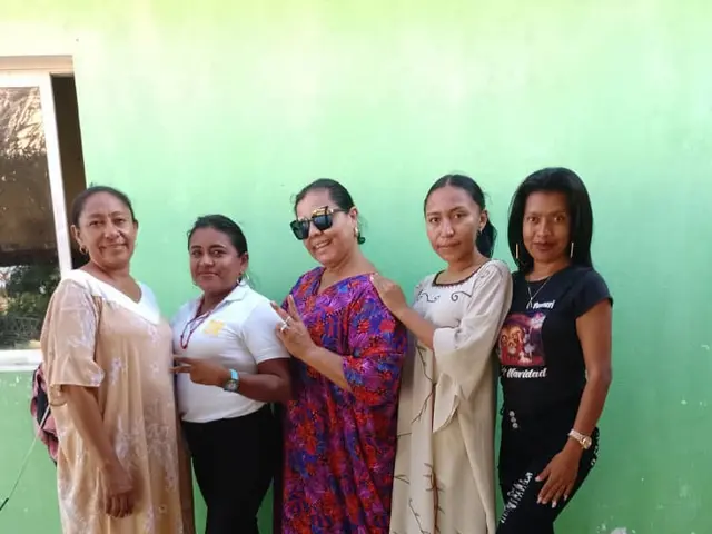 The image shows a group of women standing next to each other in front of a green wall, with a...